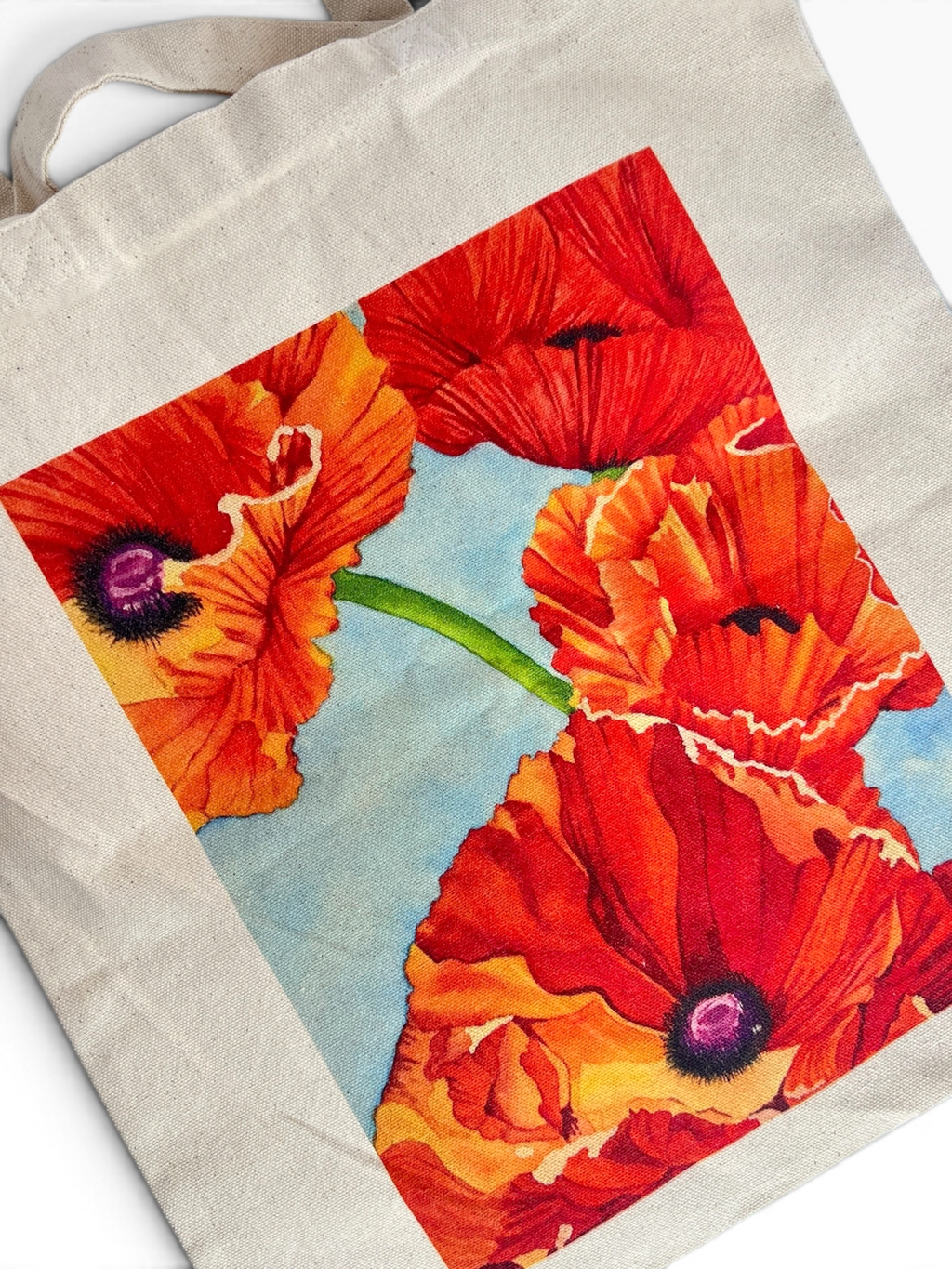 A beige canvas tote bag featuring a large, vibrant graphic print of red and orange poppies on a light blue background.