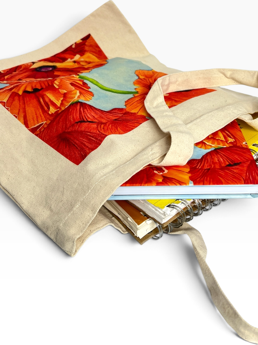 A beige canvas tote bag with a large print of red and orange poppies on the front, shown holding notebooks and books on a white background.