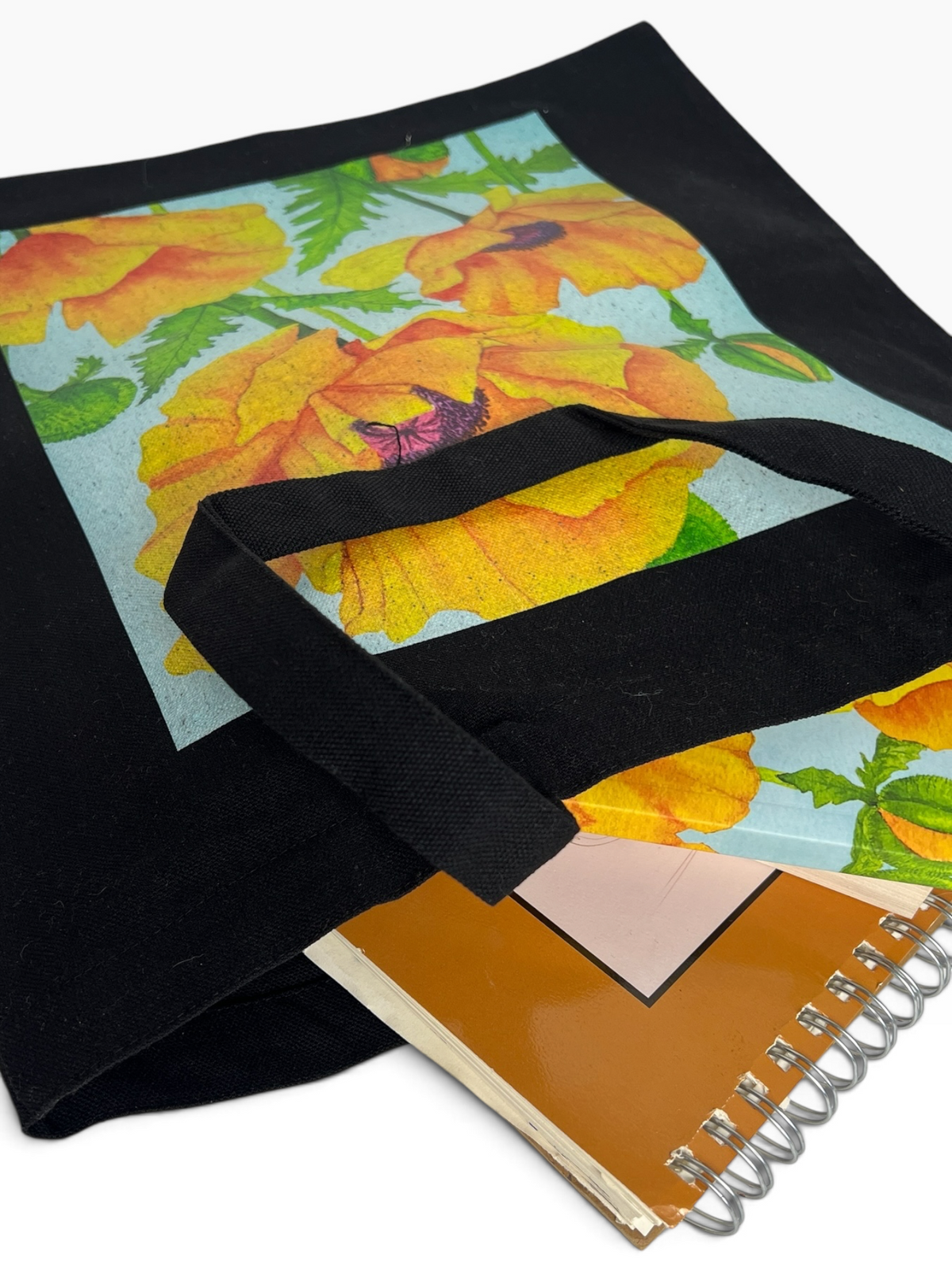 A black canvas tote bag with a light blue floral print featuring large yellow-orange poppy flowers and green leaves, shown with a spiral notebook on a white background.